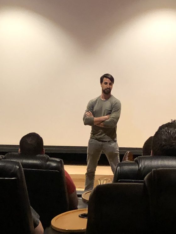 A man in a grey shirt standing in front of a screening-room audience, arms folded