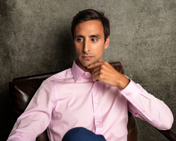 Tommy Hallal seated, chin resting on his hand, in a pink dress shirt